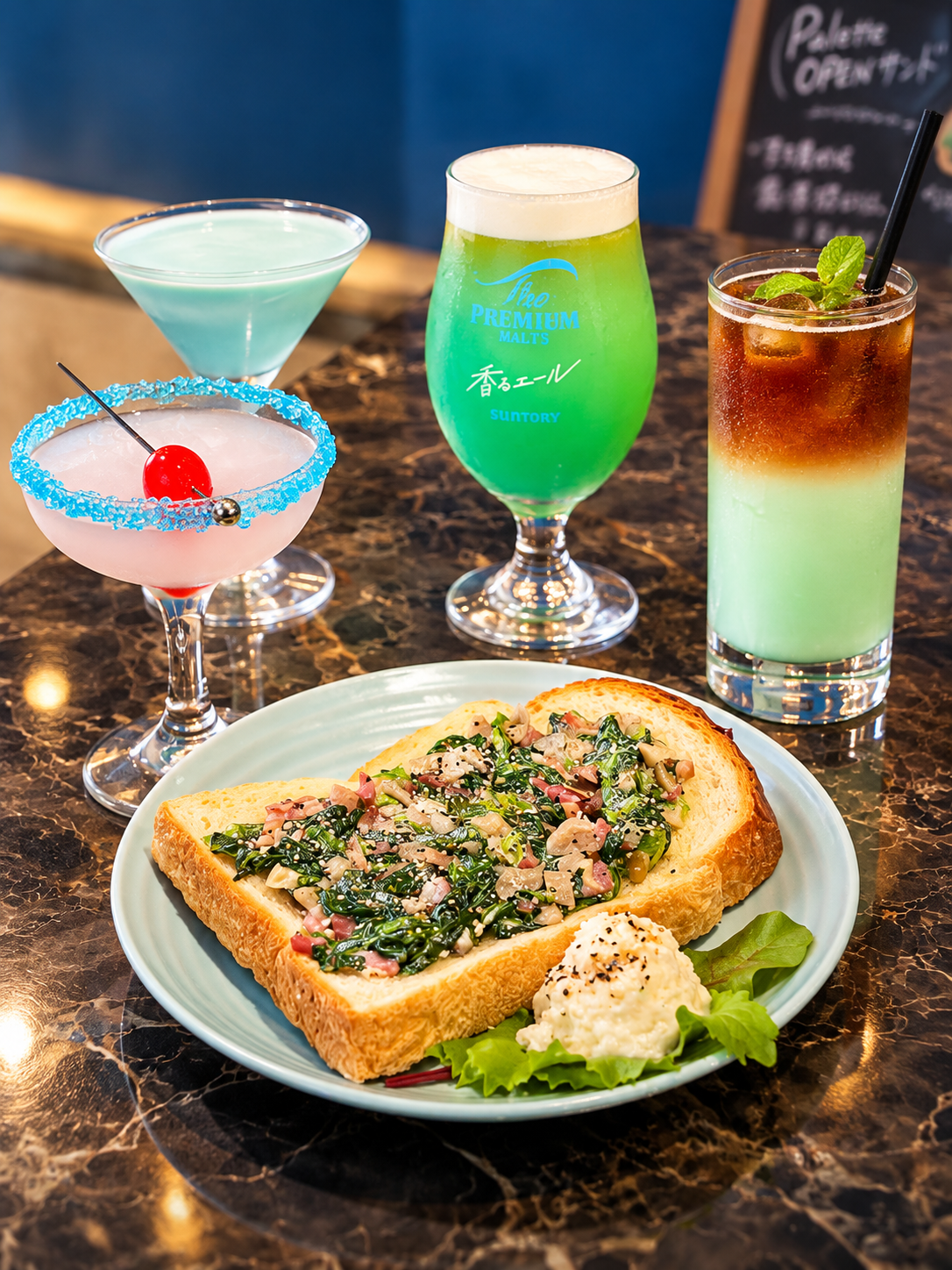 青いカクテル、ピンクのカクテル、緑のビアカクテル、アイスコーヒーと、ほうれん草のオープンサンドが大理石のテーブルに並ぶカフェメニュー