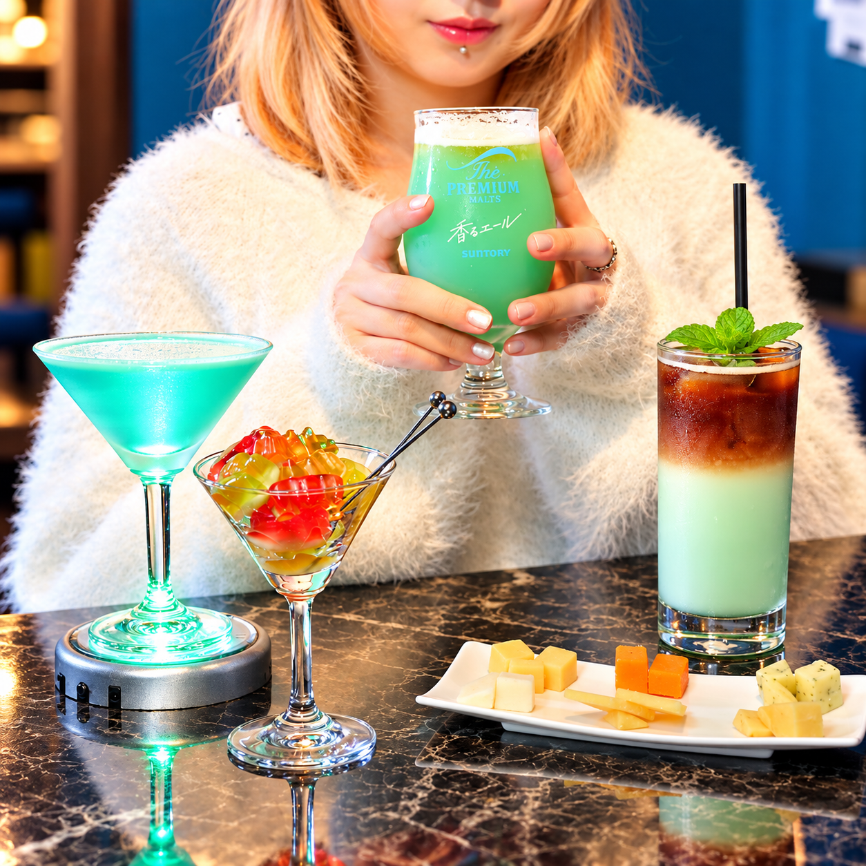 緑のビールカクテルを手にした女性と、青緑のカクテル、グミベアーを盛ったグラス、二層のアイスコーヒー、チーズの盛り合わせが黒い大理石のテーブルに並ぶ様子