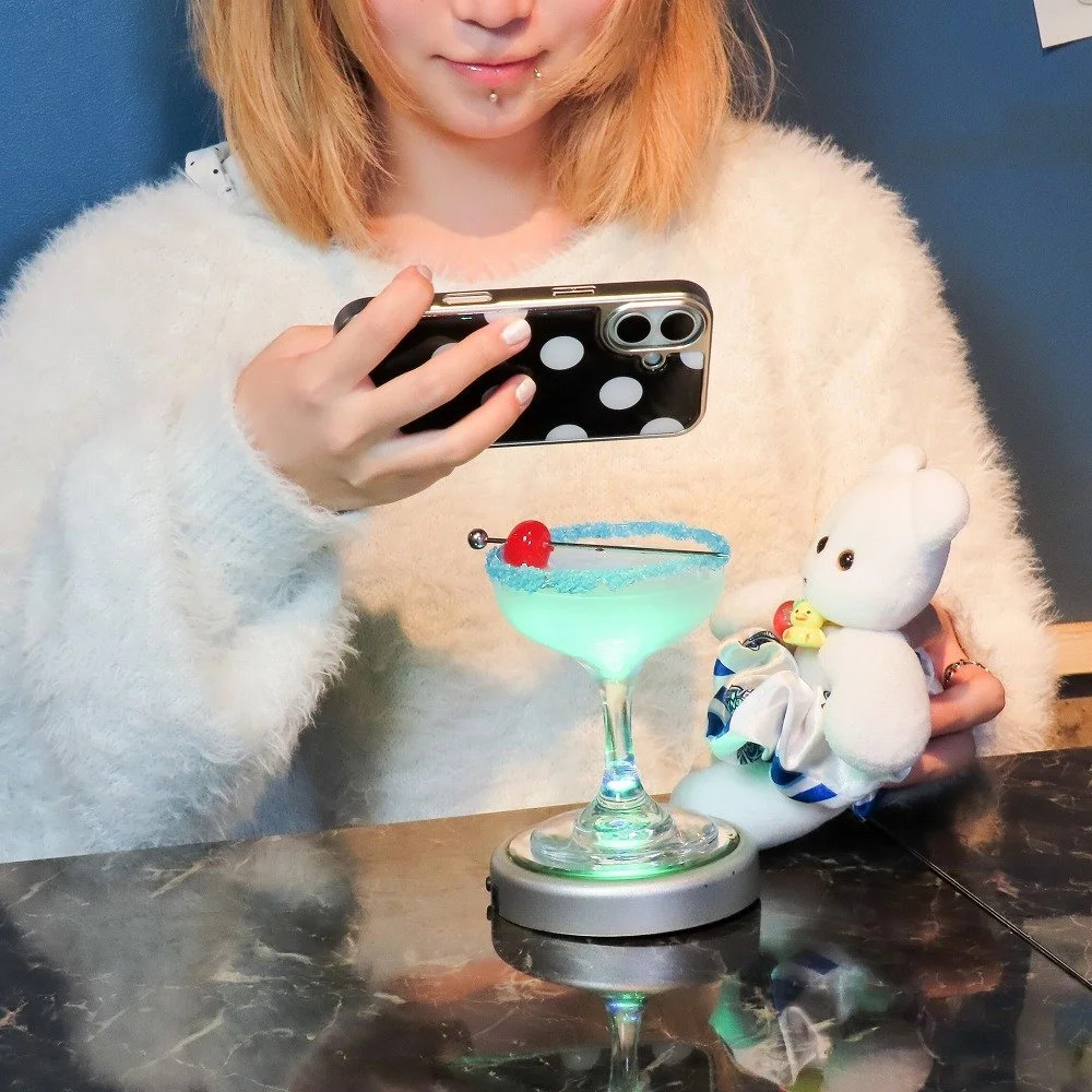 スマートフォンでドリンクを撮影する女性と、ぬいぐるみ、青く光るカクテルグラス
